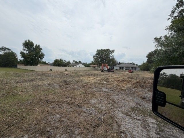 Empty gravel lot with a small tractor near houses under a cloudy sky, seen from a vehicle window