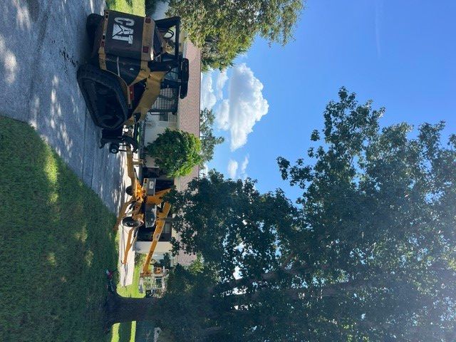 Tree-lined street with a black excavator beside a house under blue sky and clouds