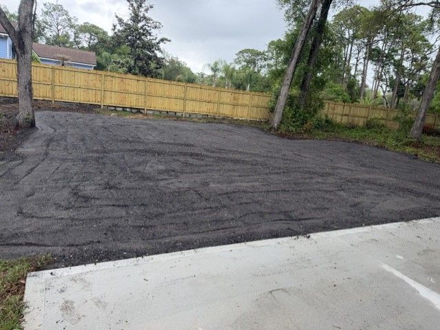 Freshly paved dark asphalt lot beside a white curb and yellow fence with trees in the background