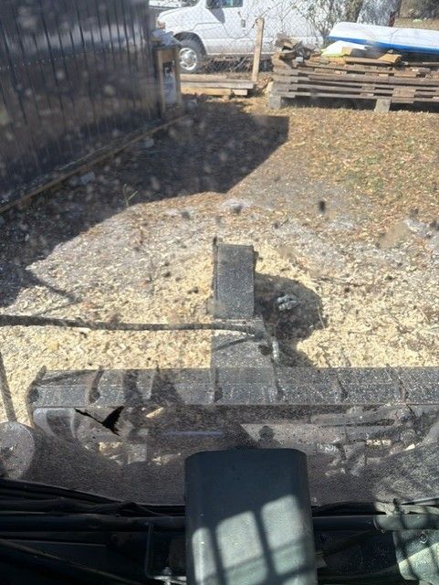 View through a vehicle windshield of a dirt yard with a black post, gravel, pallets, and a white truck in the background