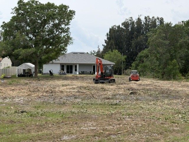 Empty lot with a house in back and two small excavators in a grassy field