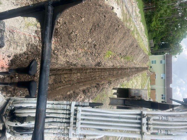 Underground utility trench with exposed pipes and cables beside a dirt path and building wall