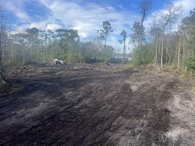 Cleared dirt lot with tire tracks beside trees under a cloudy blue sky