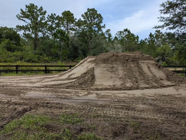 Dirt mound on a muddy dirt road beside a fence, with pine trees in the background.