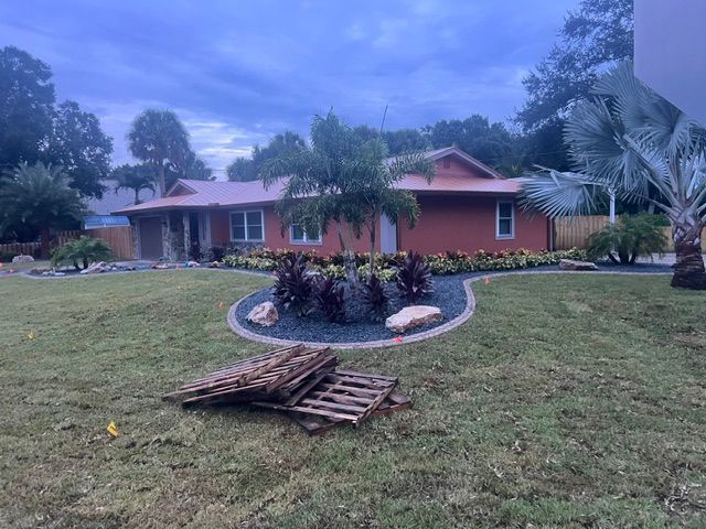 Single-story red house with palm trees, landscaped front yard, and stacked wooden pallets on the lawn.