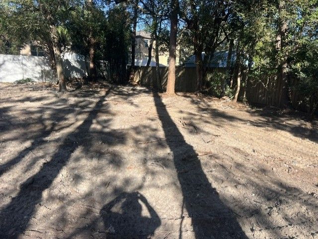 Tree-shaded dirt yard with long shadows and a fence or wall in the background