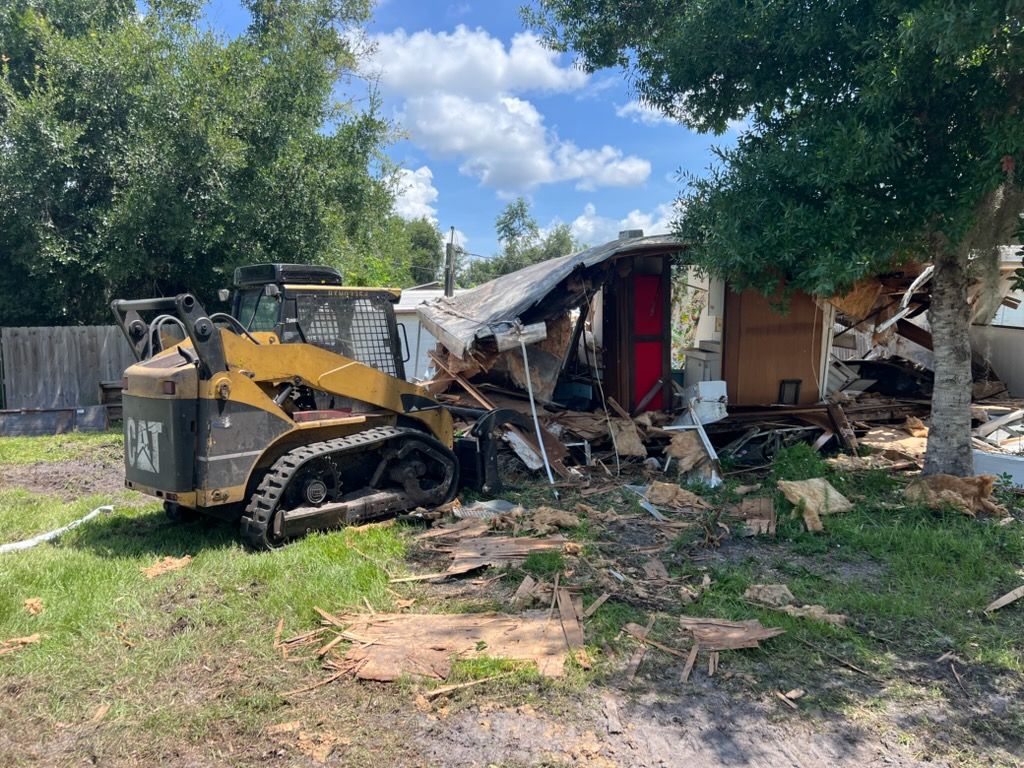 Skid steer loader demolishing a damaged house beside a tree in a yard under a blue sky