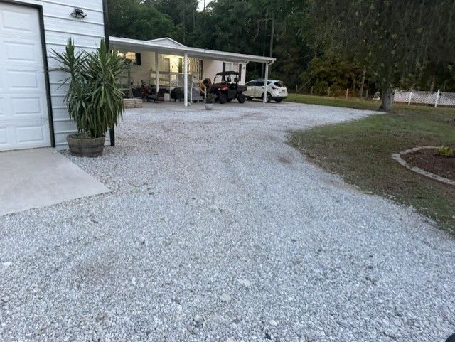 Gravel driveway leading to a lit carport beside a white house at dusk