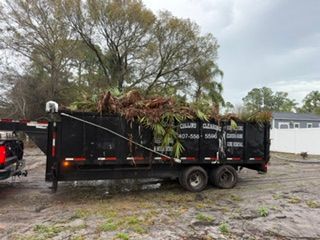 Black dump trailer loaded with tree branches in a muddy yard, with a pickup truck nearby.