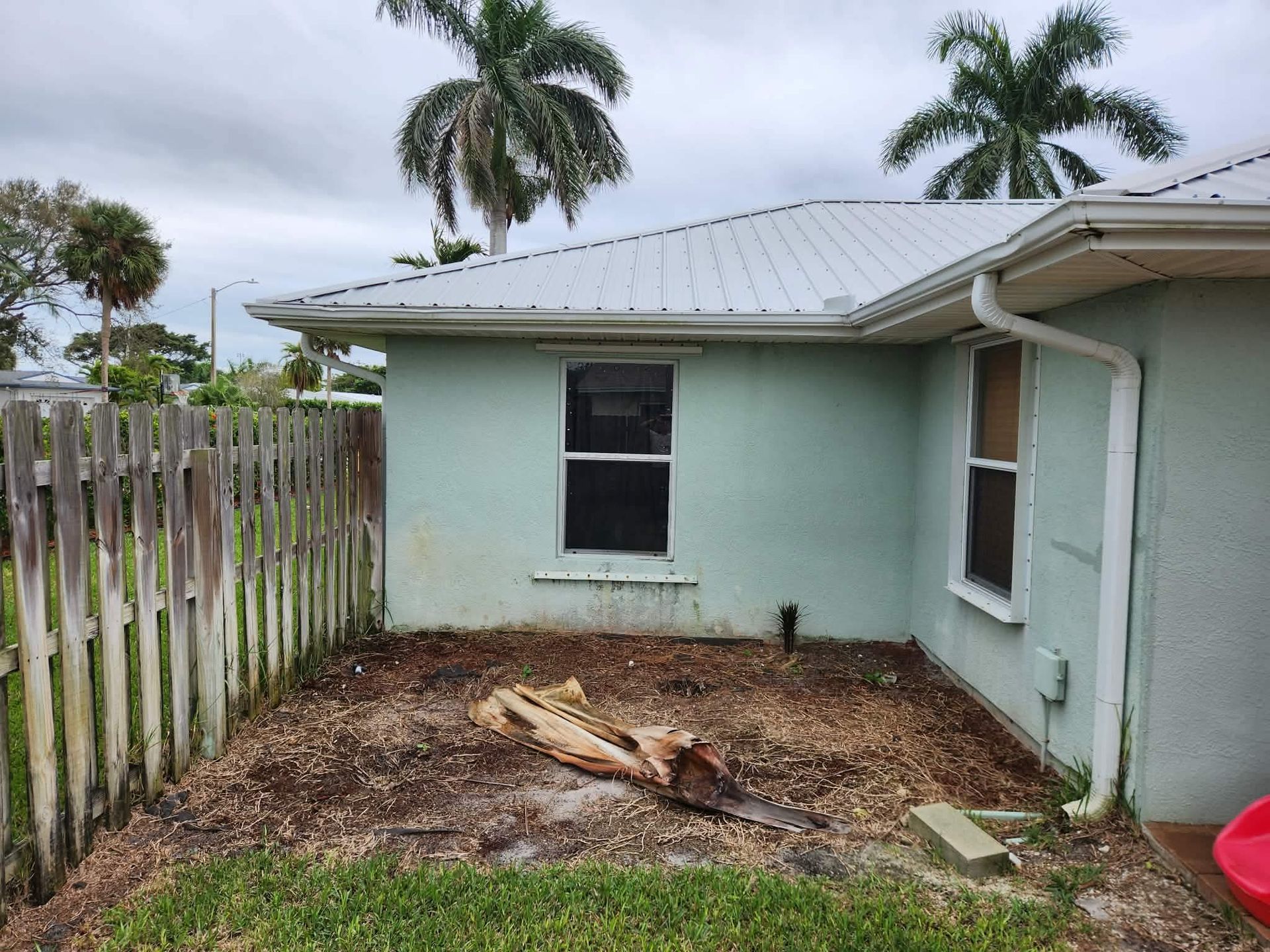 Backyard with a mint-green house wall, fence, palm trees, and a bare patch of soil and debris