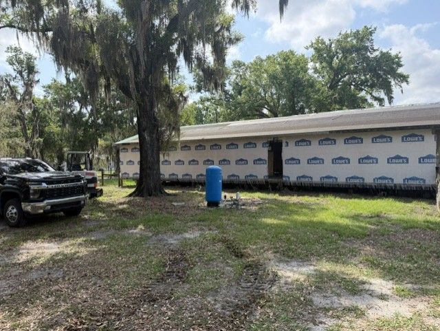 A long white building with blue trim under trees, with a black truck and blue barrel in the foreground.