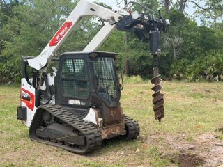 Compact tracked digger with auger attachment on grassy ground near trees