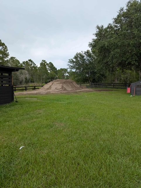 Grassy field with a dirt mound under cloudy skies, trees in the background, and a small shed at left.