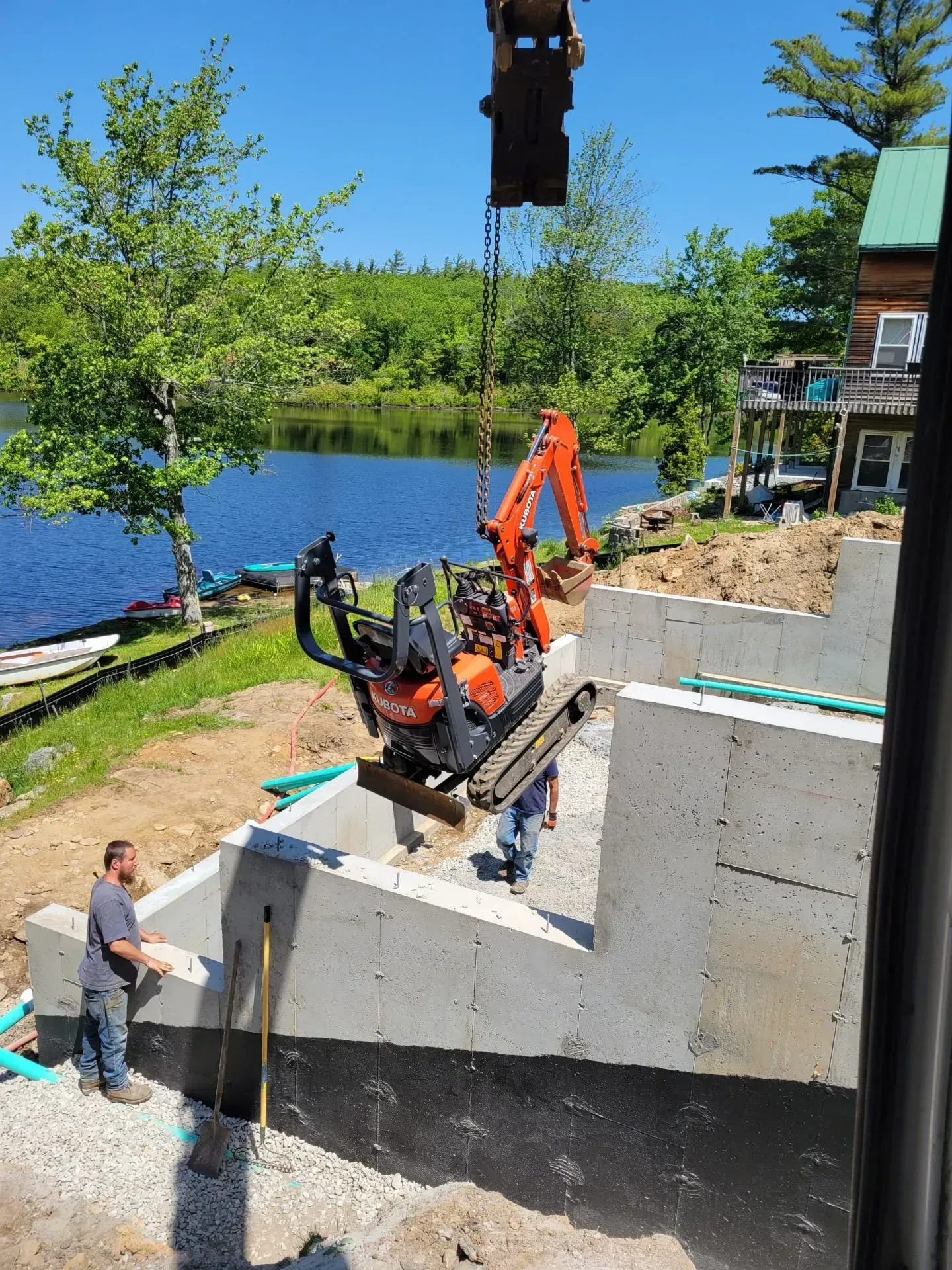 Excavator lowering a concrete wall section beside a lakeside construction site with workers nearby