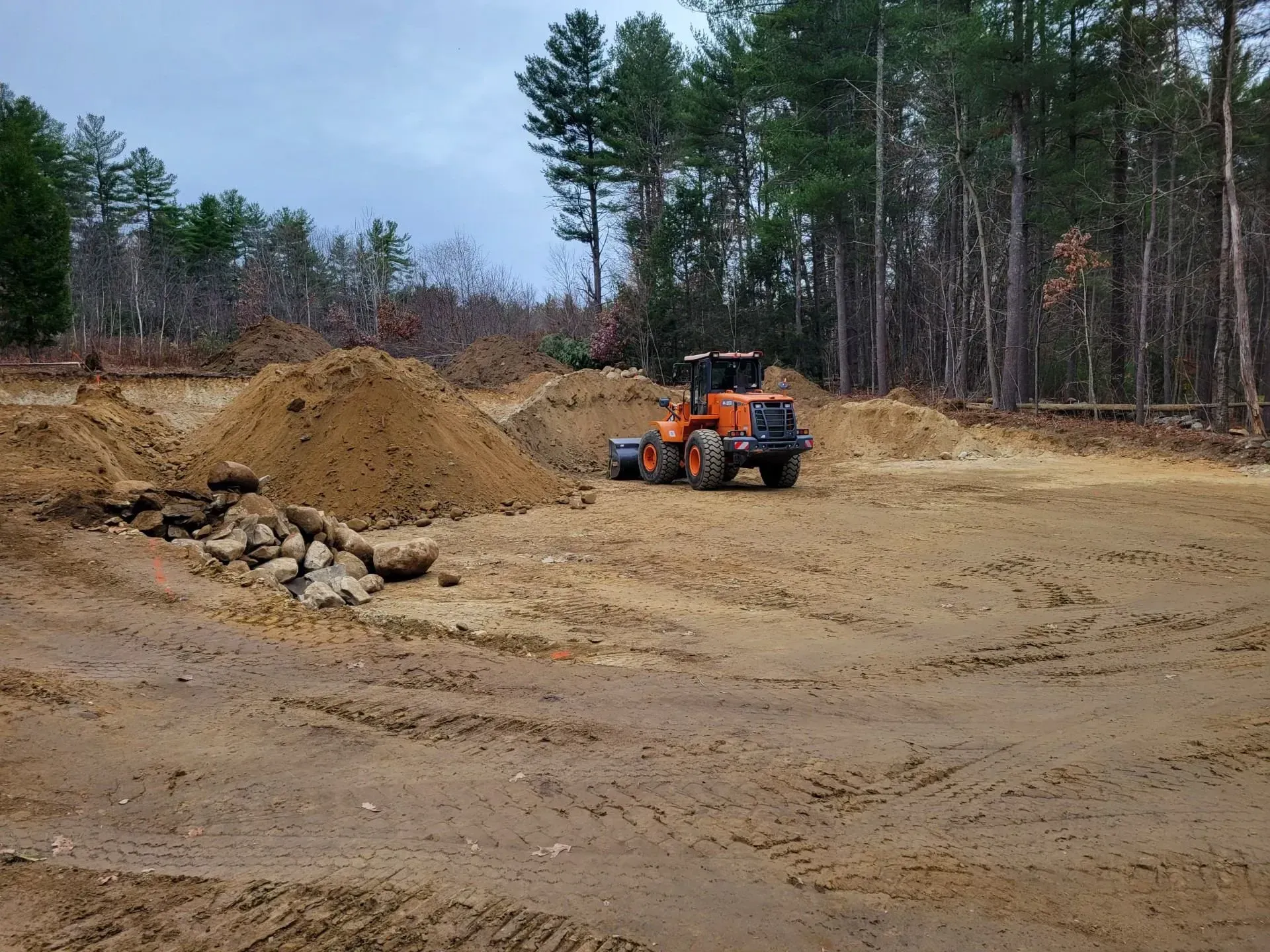 Orange loader parked on a dirt construction site beside a pile of gravel, with trees in the background