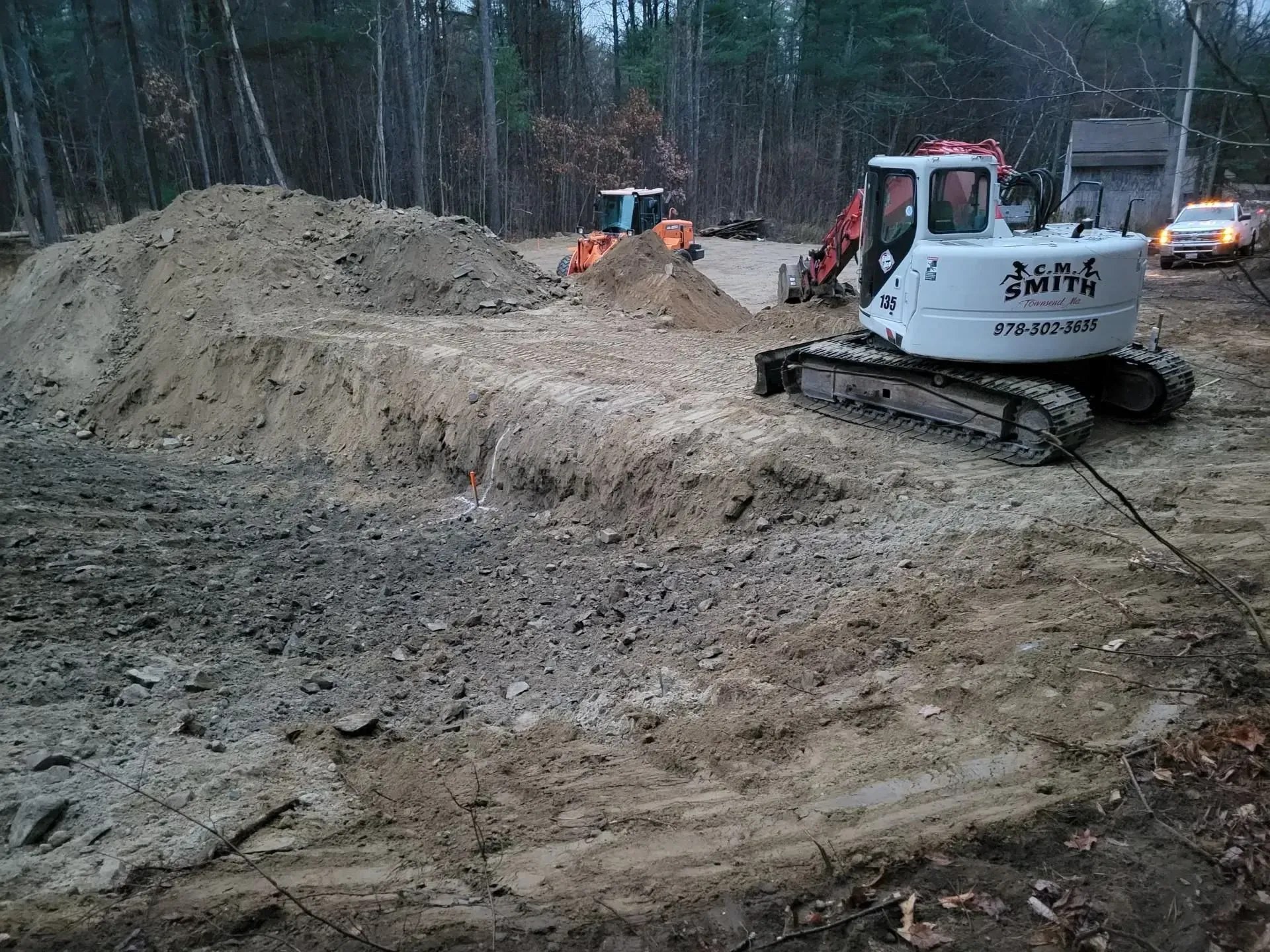 Construction site with a white tracked excavator beside a large dirt pile in a muddy wooded area.