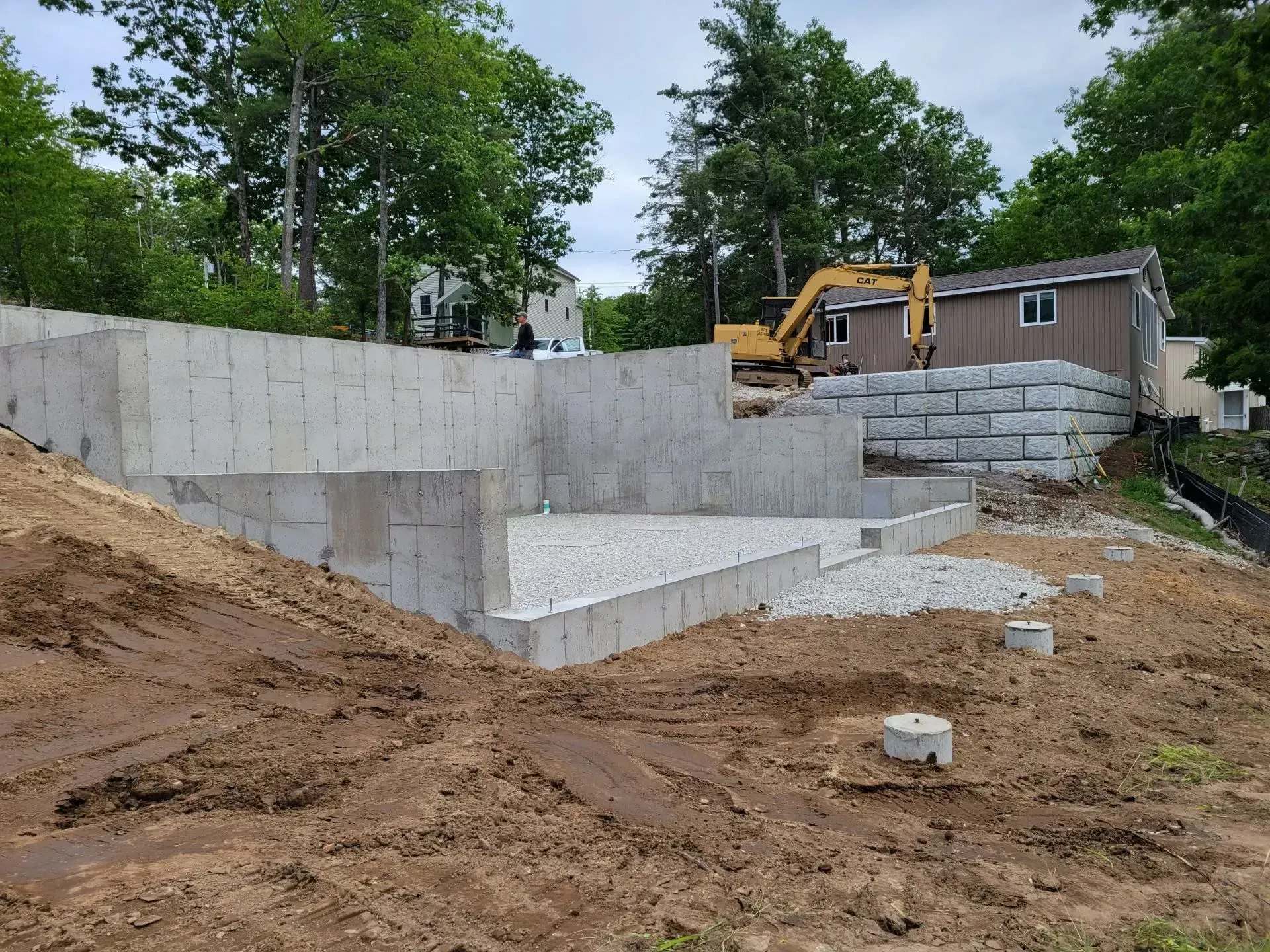 Concrete retaining wall under construction beside a house, with an excavator and piles of dirt on a sloped site
