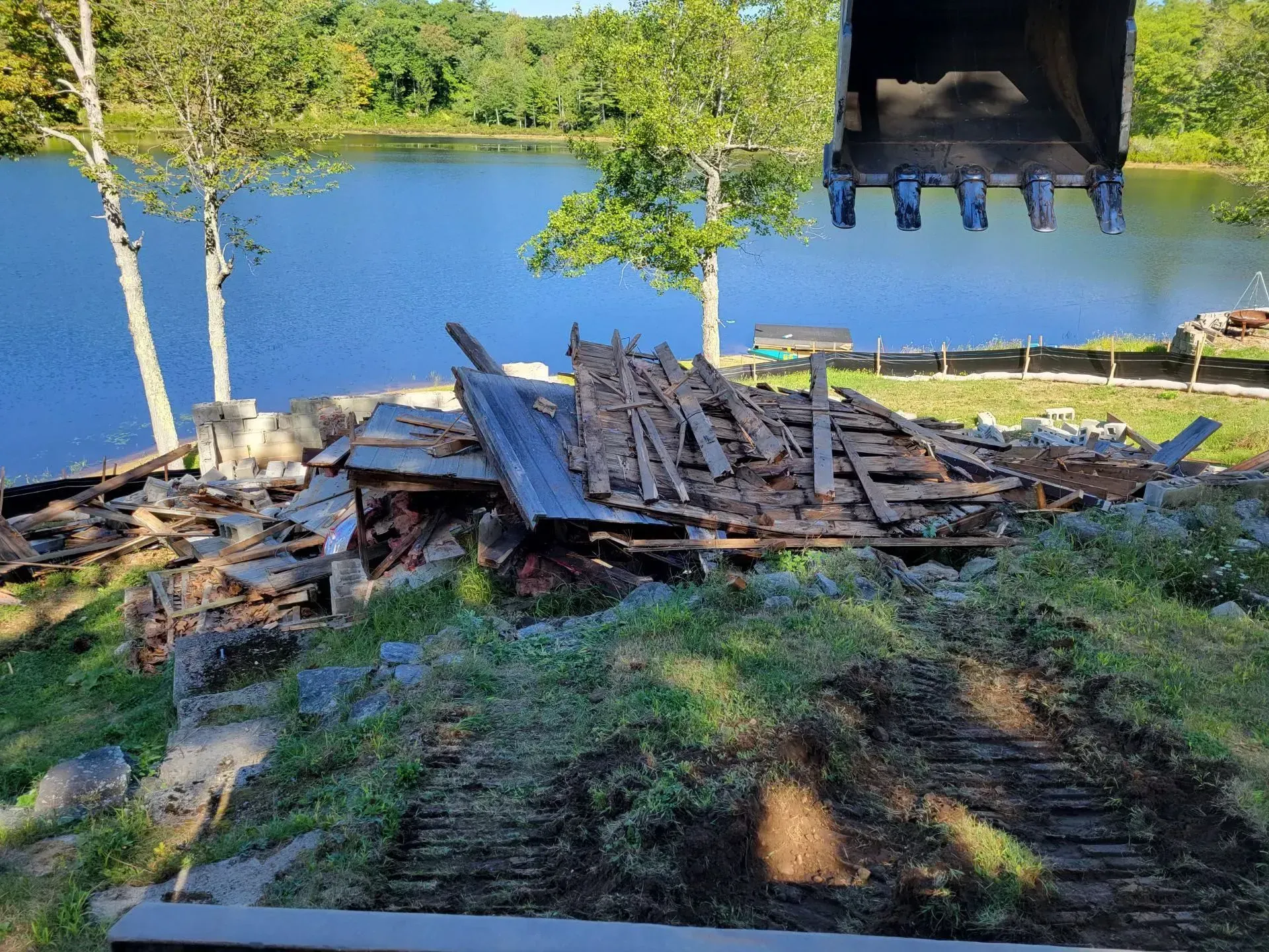 Collapsed wooden debris by a lakeshore under a bridge, with trees and water in the background.