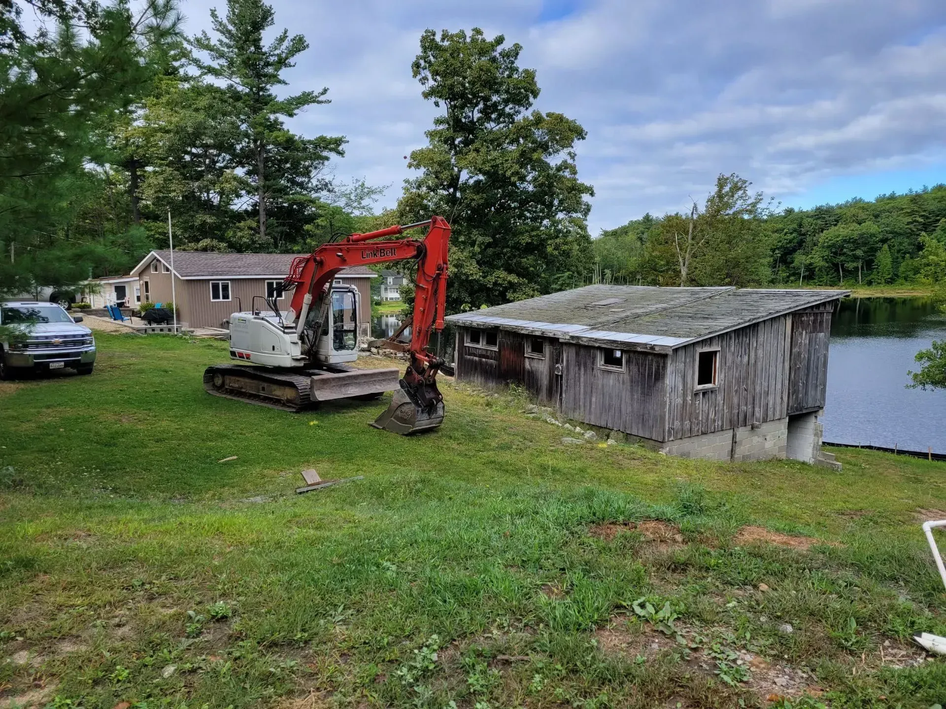 Excavator beside a weathered lakeside shed in a grassy yard with trees and a parked car