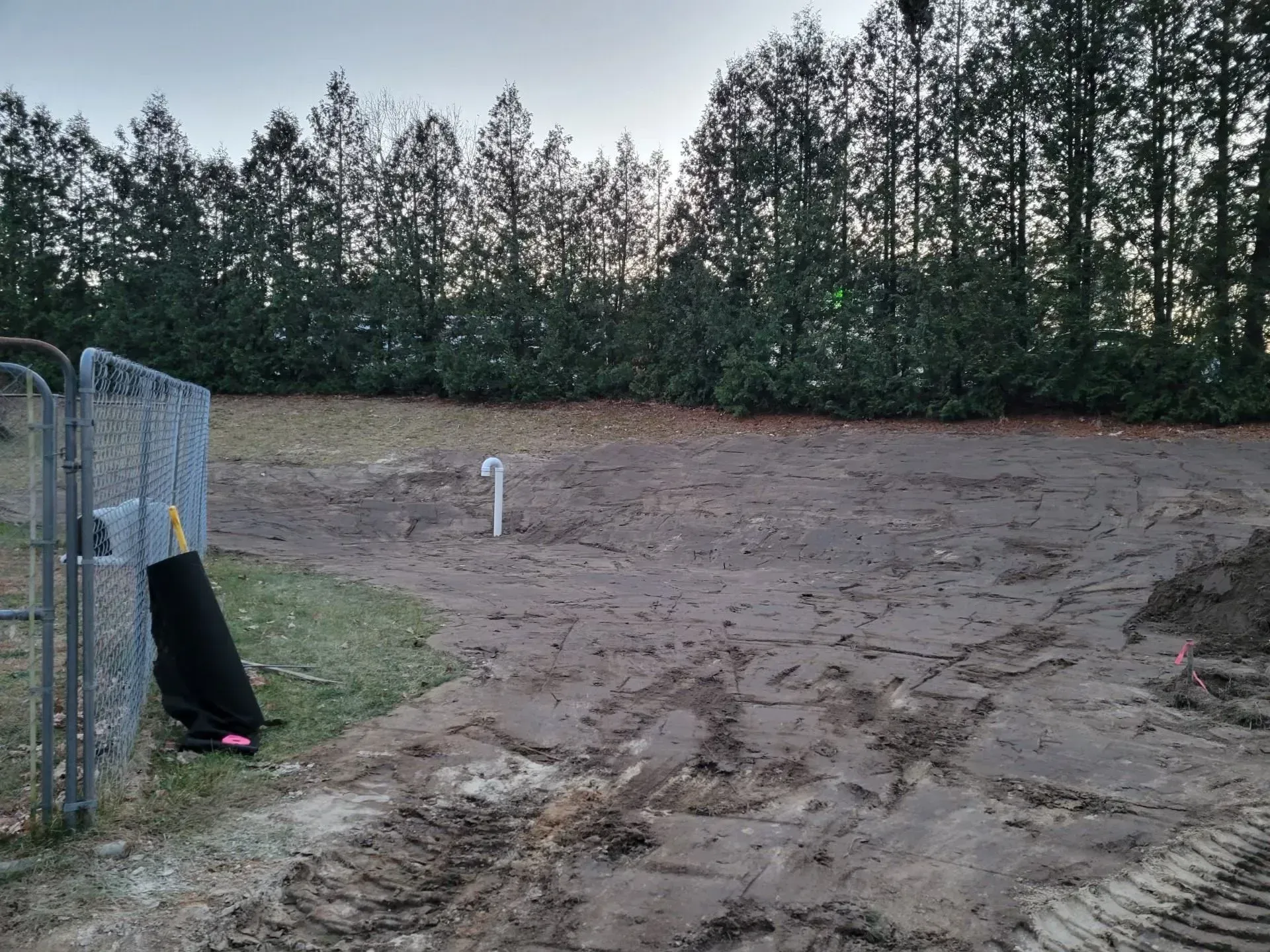 Dirt clearing beside a fence and trees, with tire tracks and a black tarp on the left.