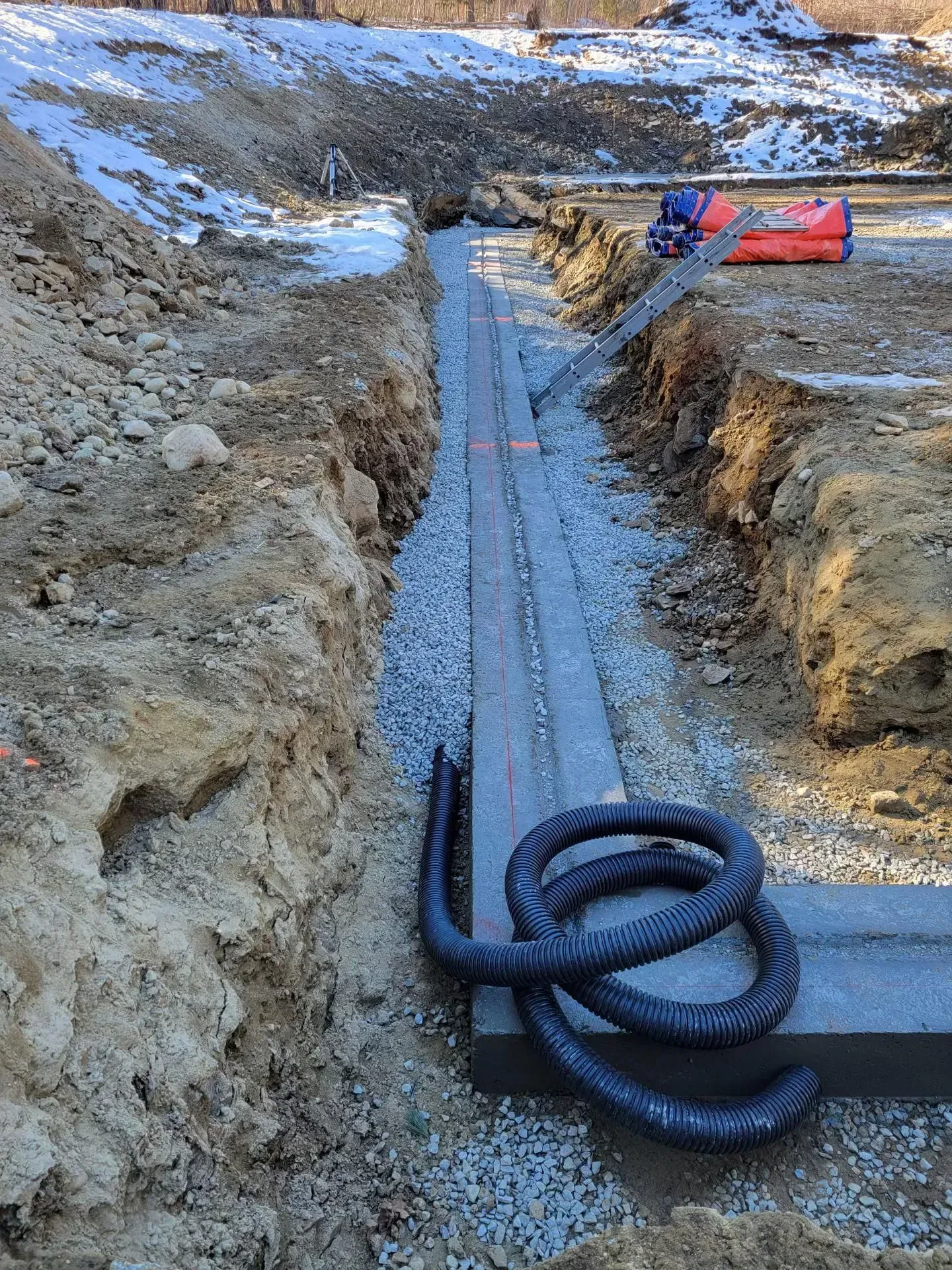 Trench with coiled black pipe laid in gravel, workers in the distance near an orange barrier.