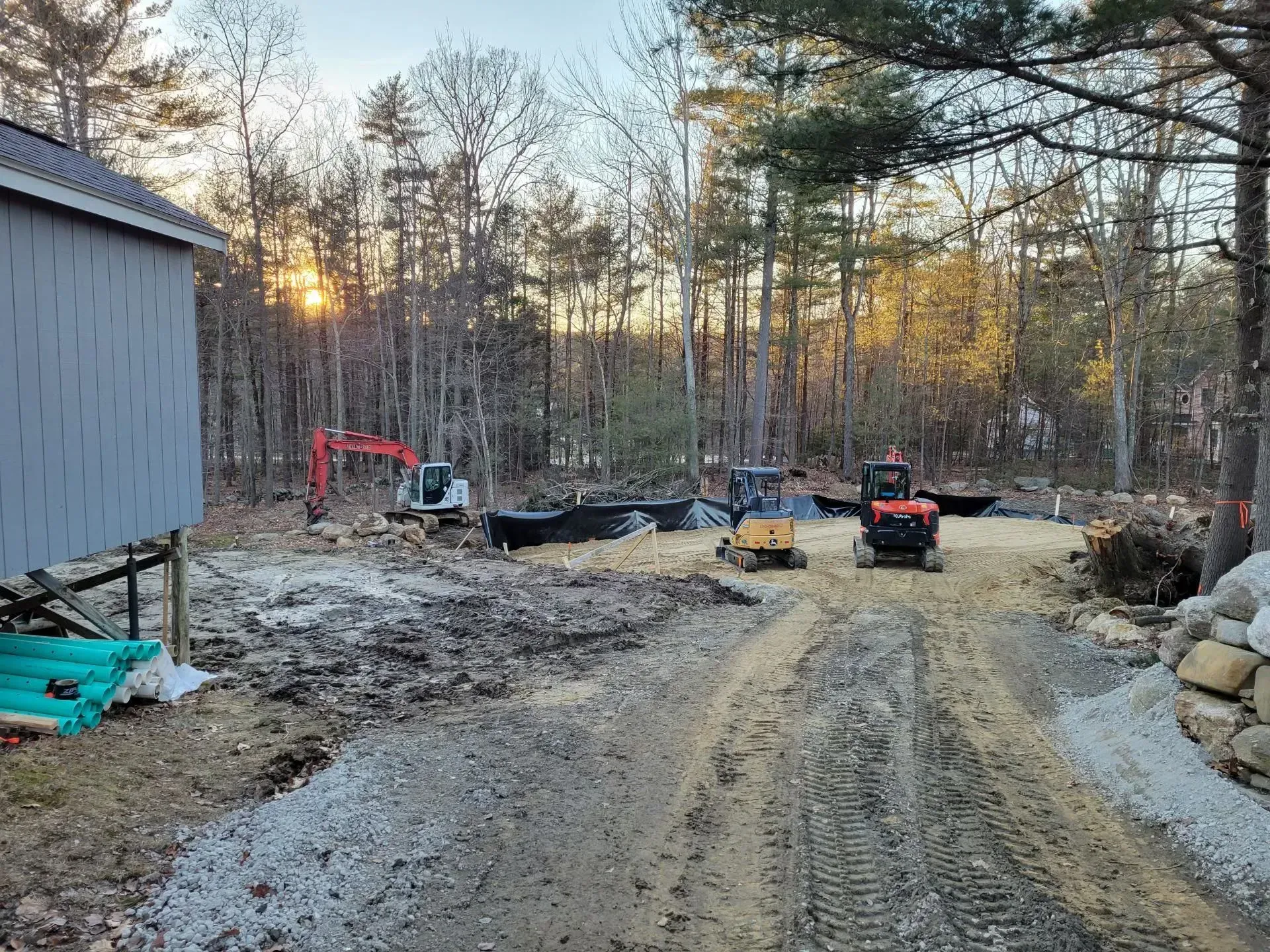 Construction site with dirt road, excavator, utility vehicle, and snow-dusted ground beside a building