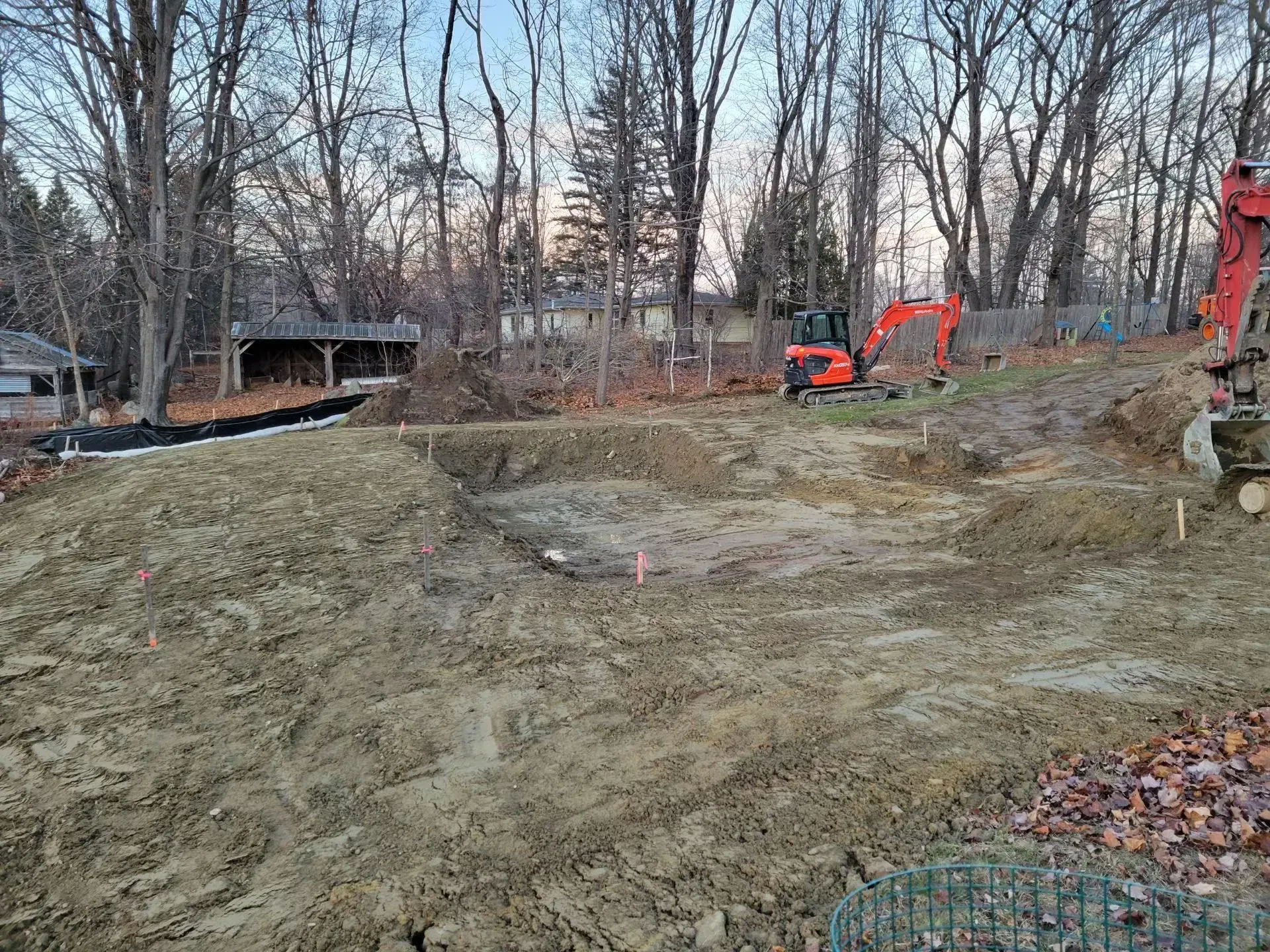 Excavator grading a bare dirt lot beside trees and a house at dusk.