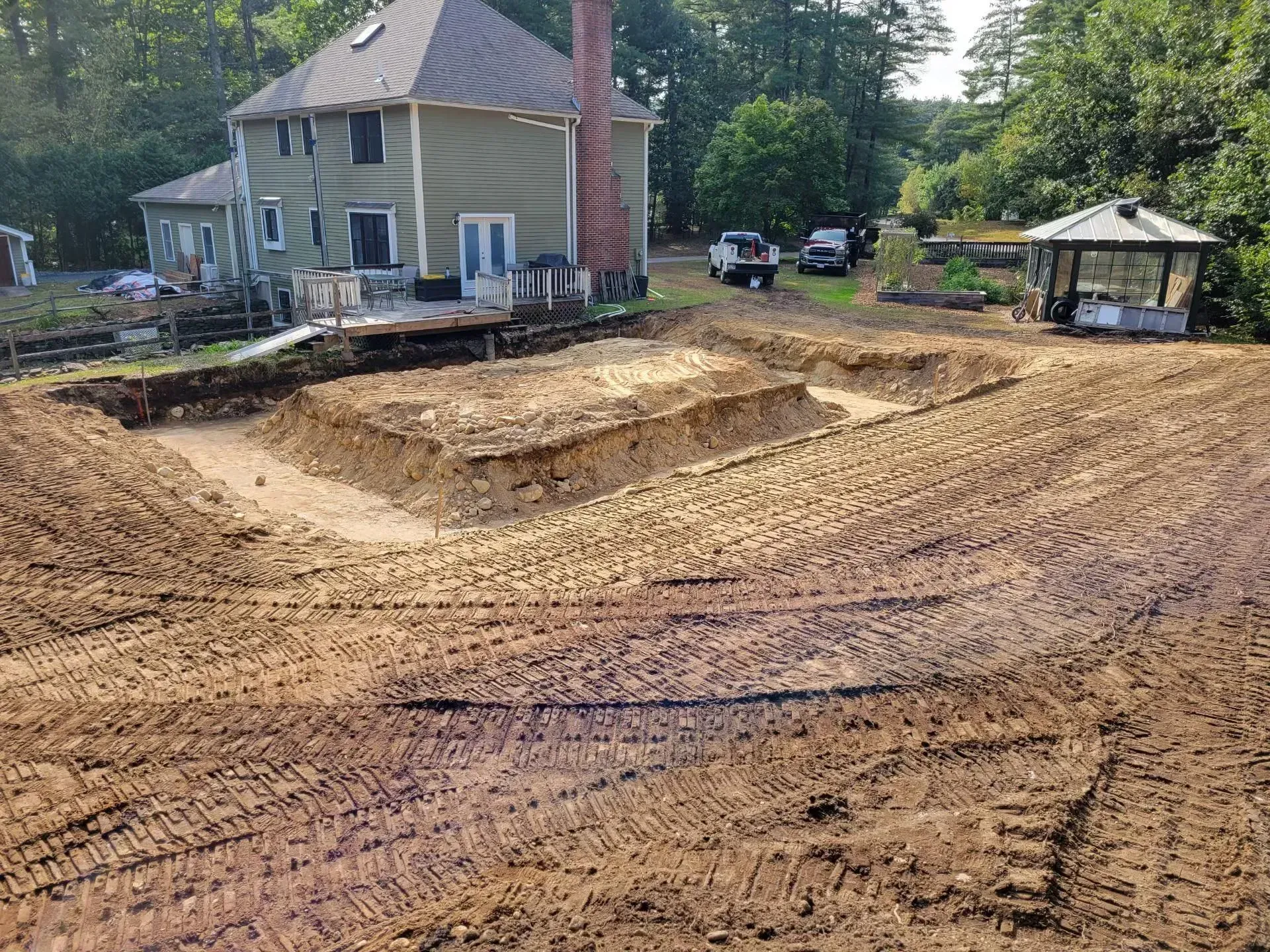 Excavated backyard with dirt berms beside a house, surrounded by trees and a gazebo