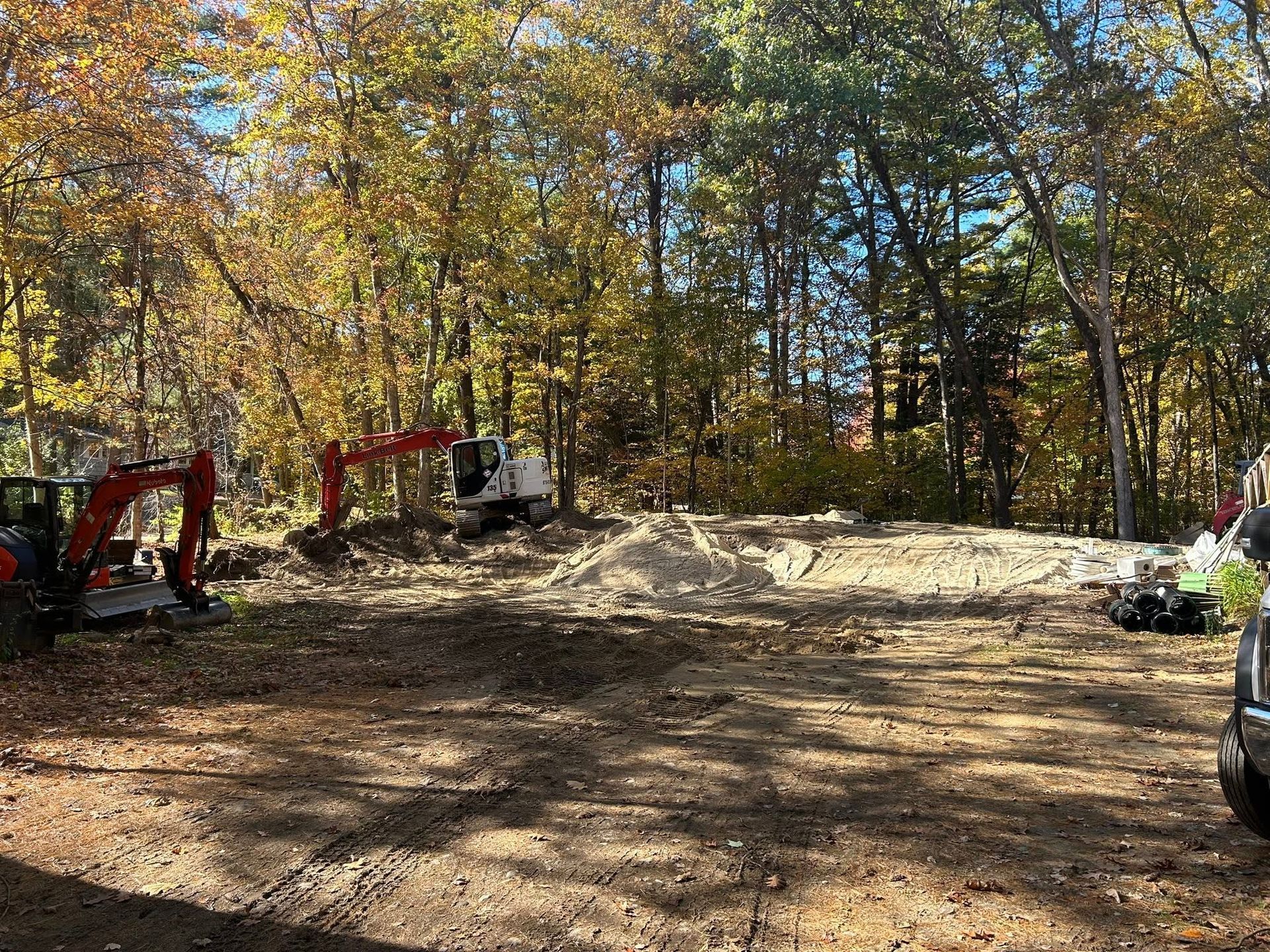 Excavators and trucks clearing a dirt lot beside a wooded area in autumn.