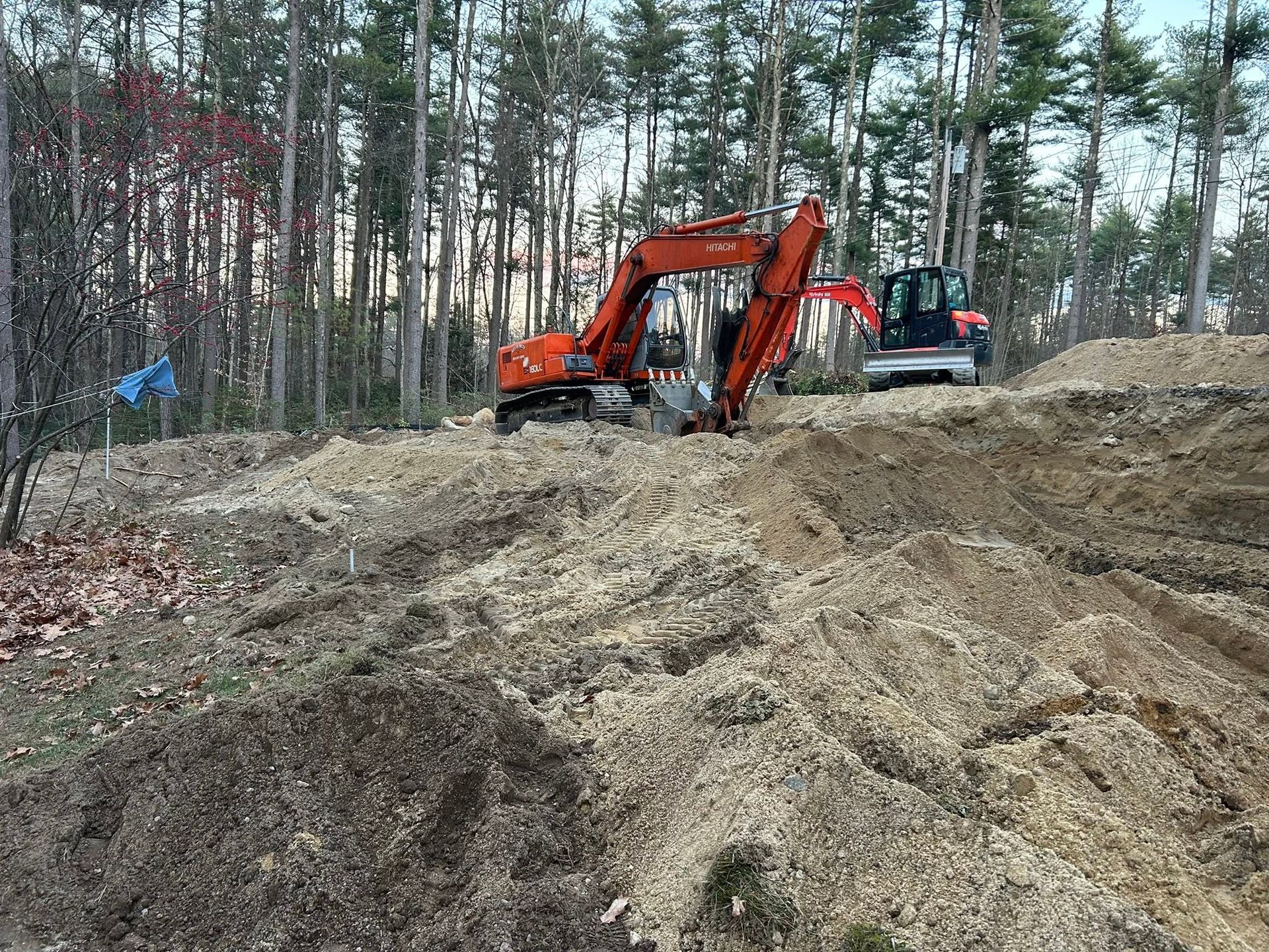 Orange excavator on a dirt mound at a forest work site, with a red truck in the background.
