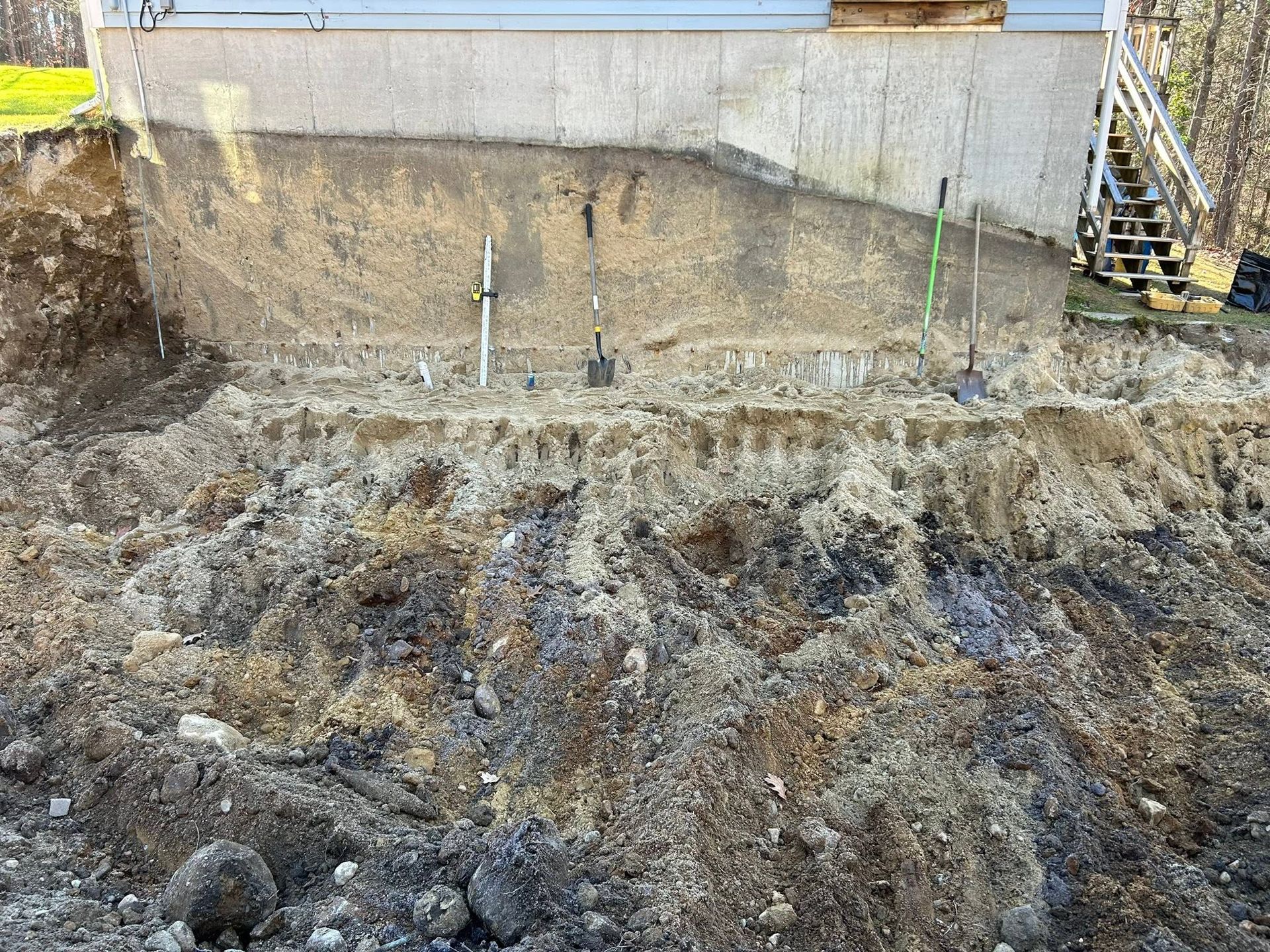 A large excavated dirt slope beside a concrete retaining wall with survey stakes and a stairway on the right.