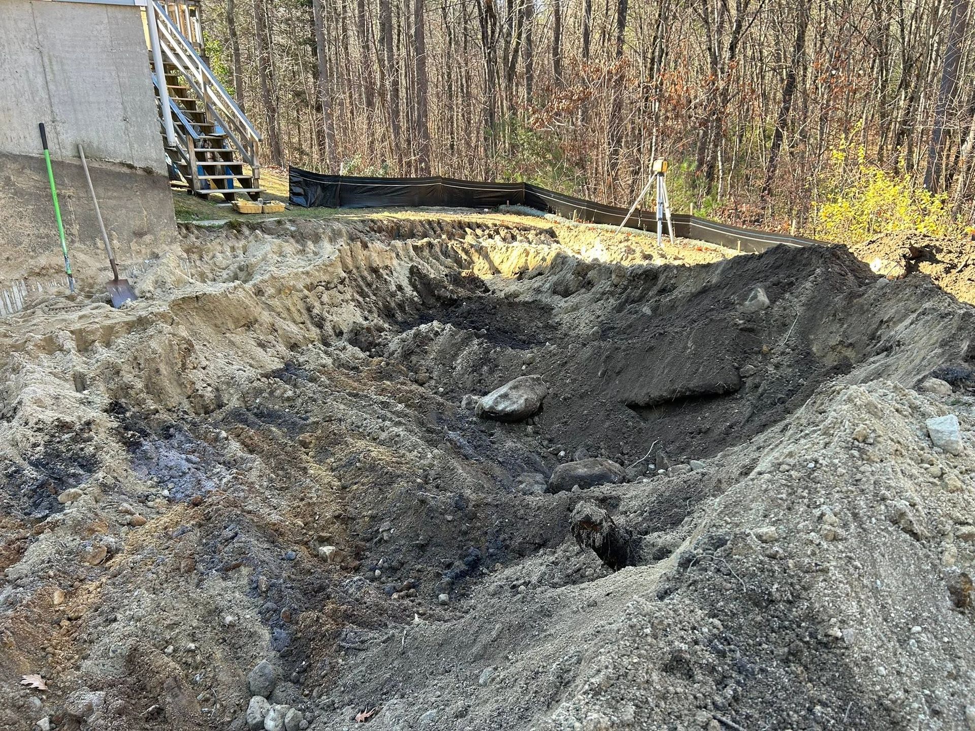 Large dirt excavation pit beside a building with stairs, surrounded by trees and bare ground.
