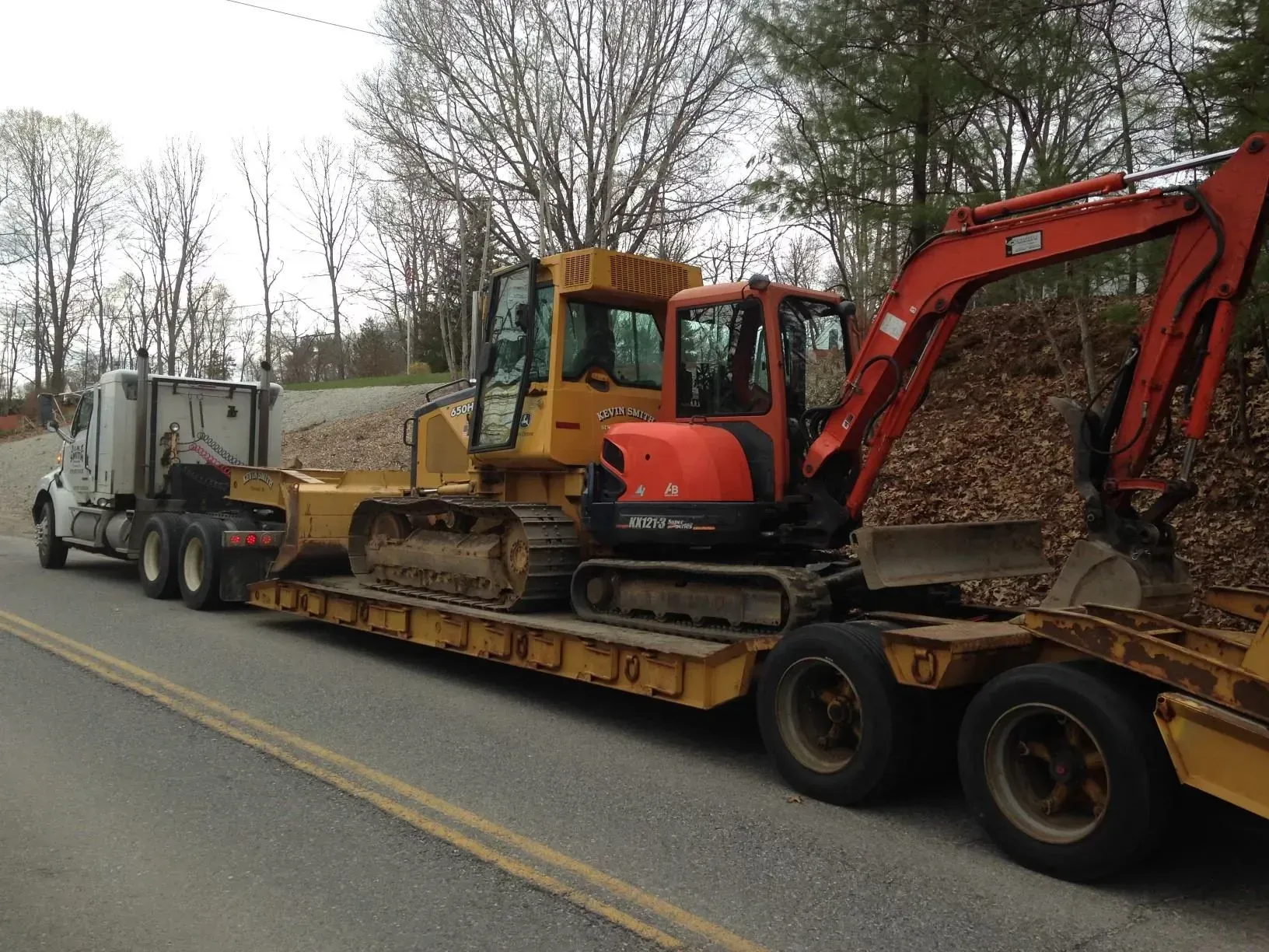 Trailer hauling a tracked excavator and dump truck along a roadside in a wooded area