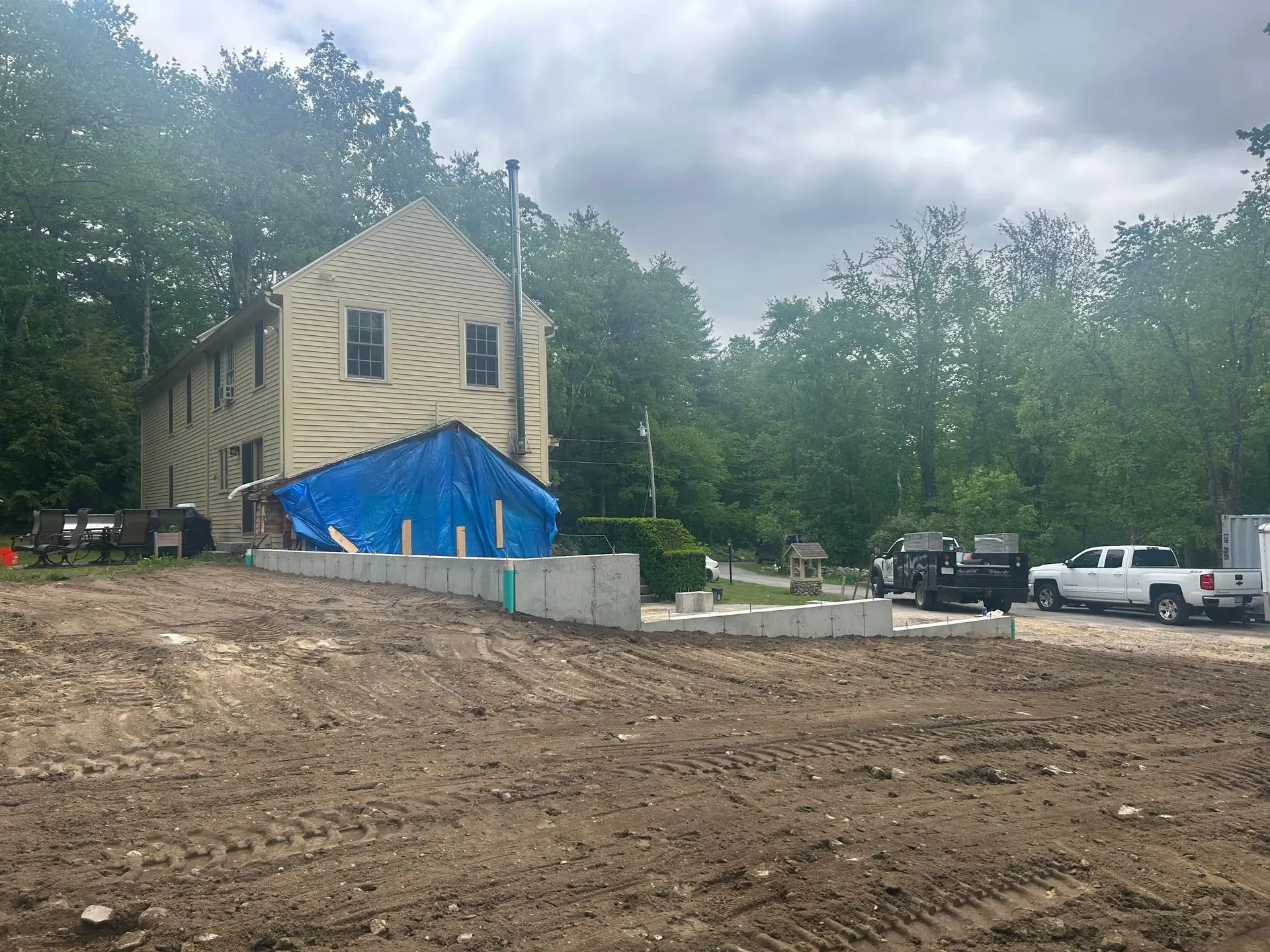 Construction site with partially built house, blue tarp, dirt foreground, and parked trucks under cloudy sky