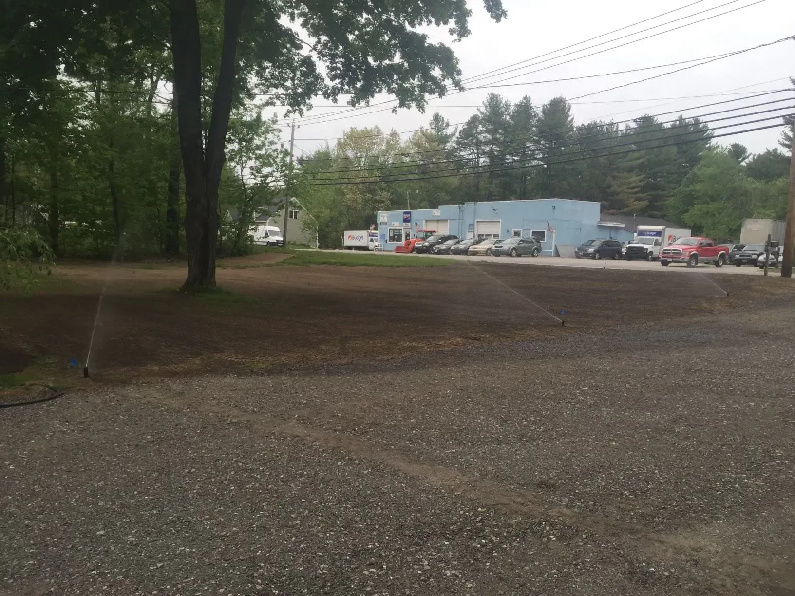 Gravel lot with trees and a blue building in the background, with several parked vehicles under an overcast sky