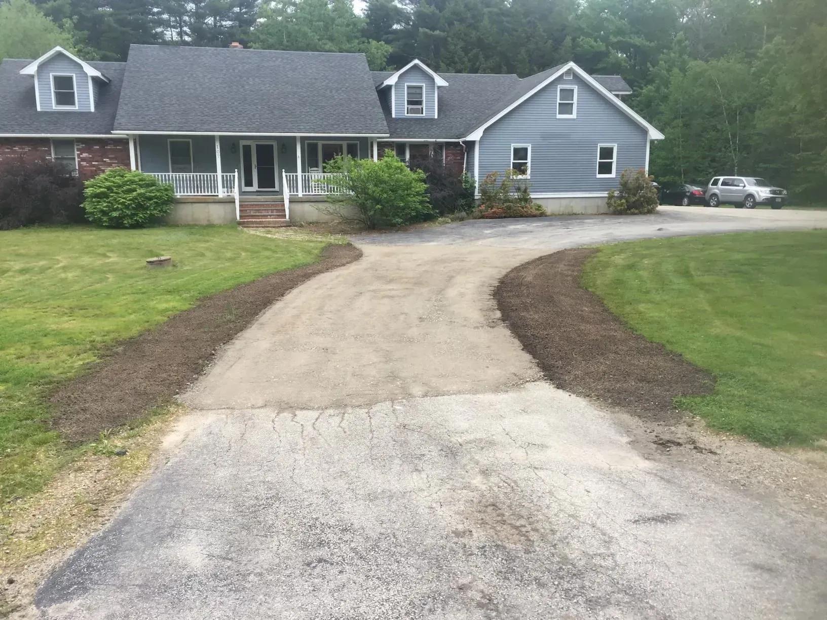 Curved driveway leading to a gray house with front porch and attached garage, bordered by green lawn and trees.
