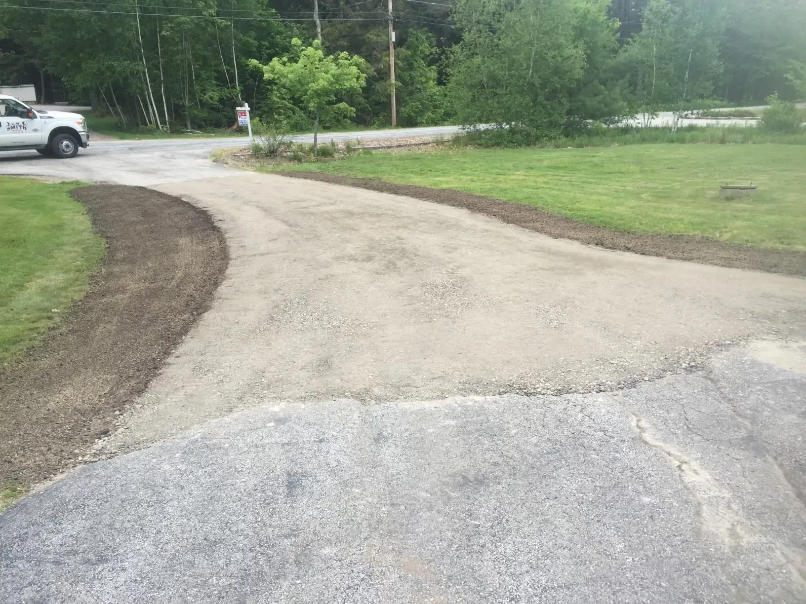 Curving gravel driveway leading through a green wooded area, with a parked white truck at left.