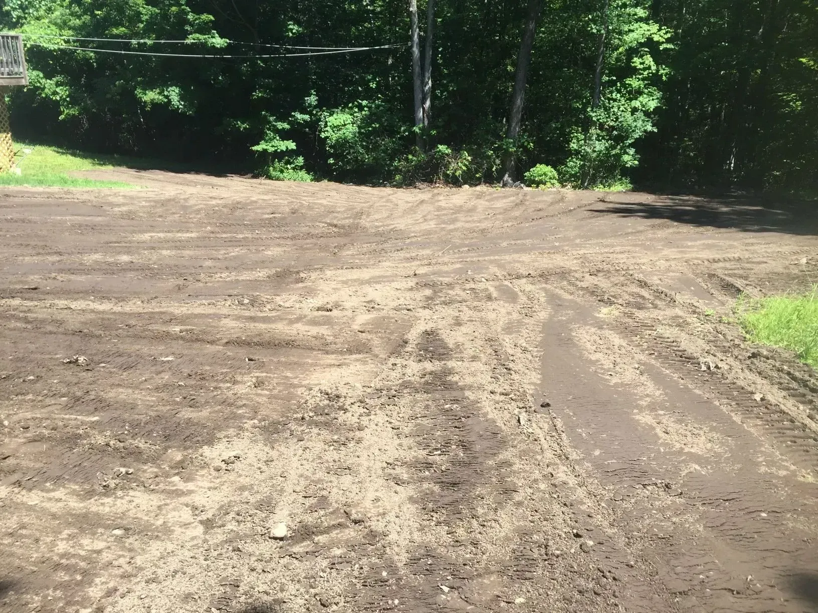 Cleared dirt lot with tire tracks beside dense green trees and brush