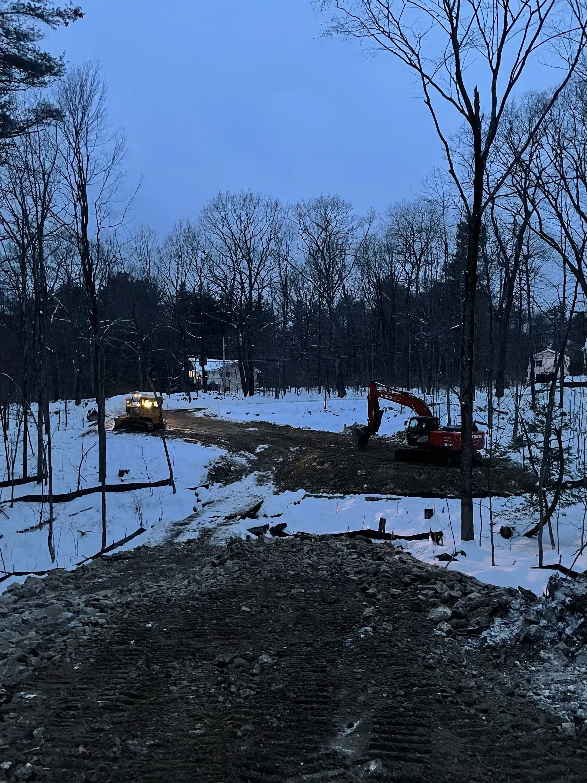 Snowy wooded site with a bulldozer at dusk and a muddy path in the foreground