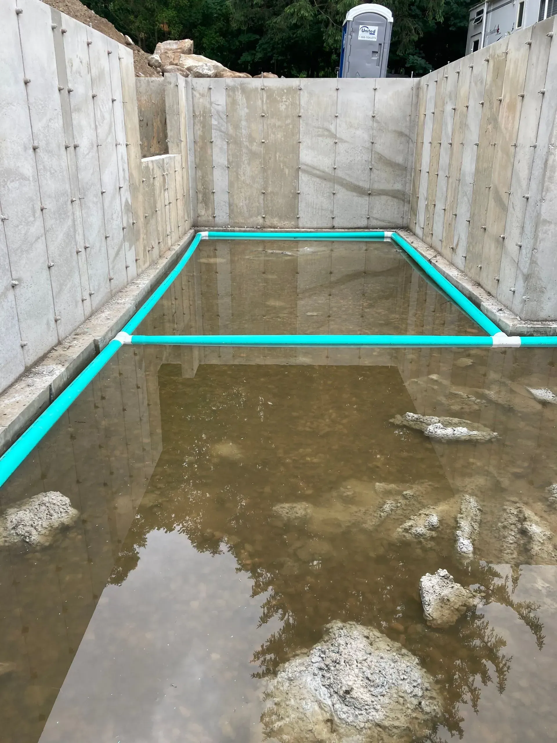 Concrete basement water pit with muddy standing water and turquoise PVC pipes along the walls and floor.