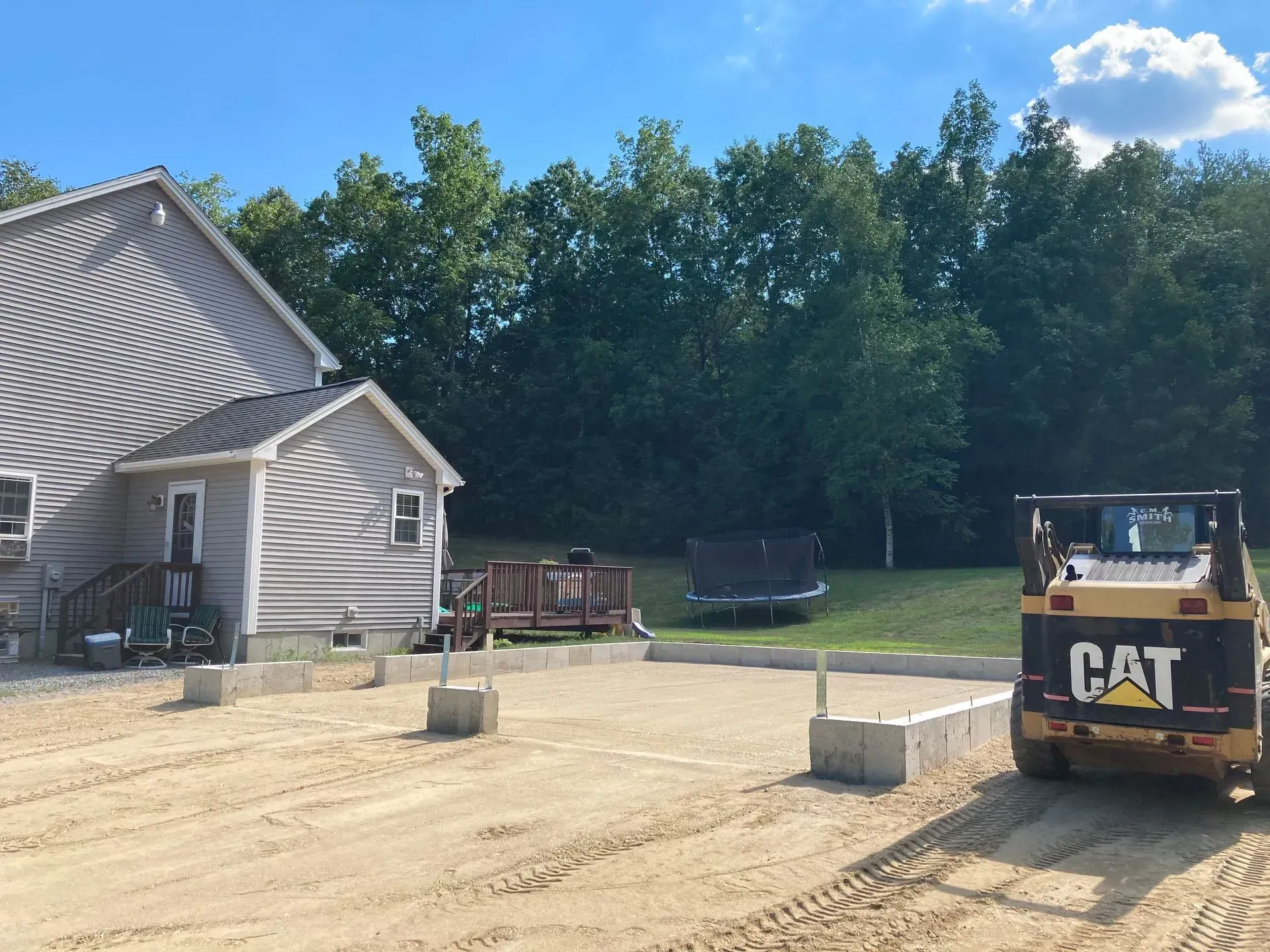 Construction site beside a gray house, with a CAT skid steer and concrete blocks on a dirt driveway.