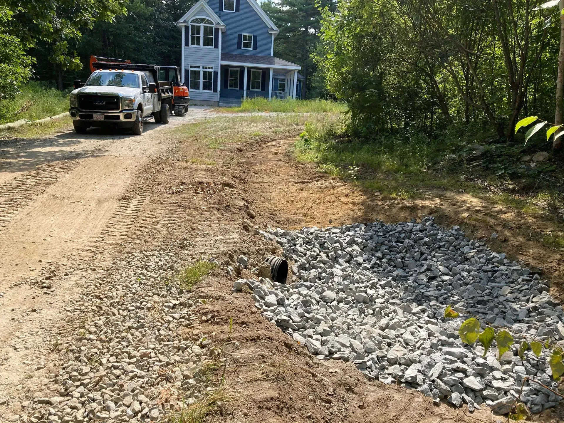 Gravel driveway beside a blue house, with parked vehicles and a rock drainage bed in the foreground