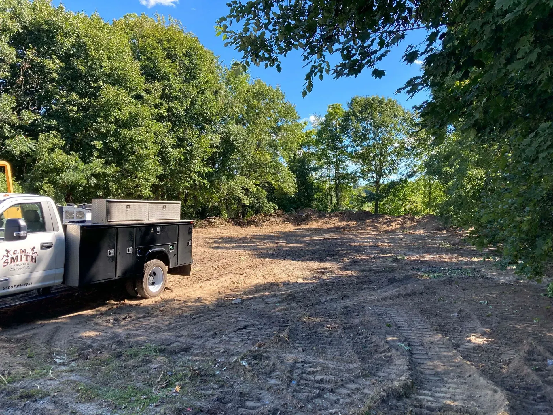 White utility truck beside a cleared dirt path in a wooded area under a bright blue sky