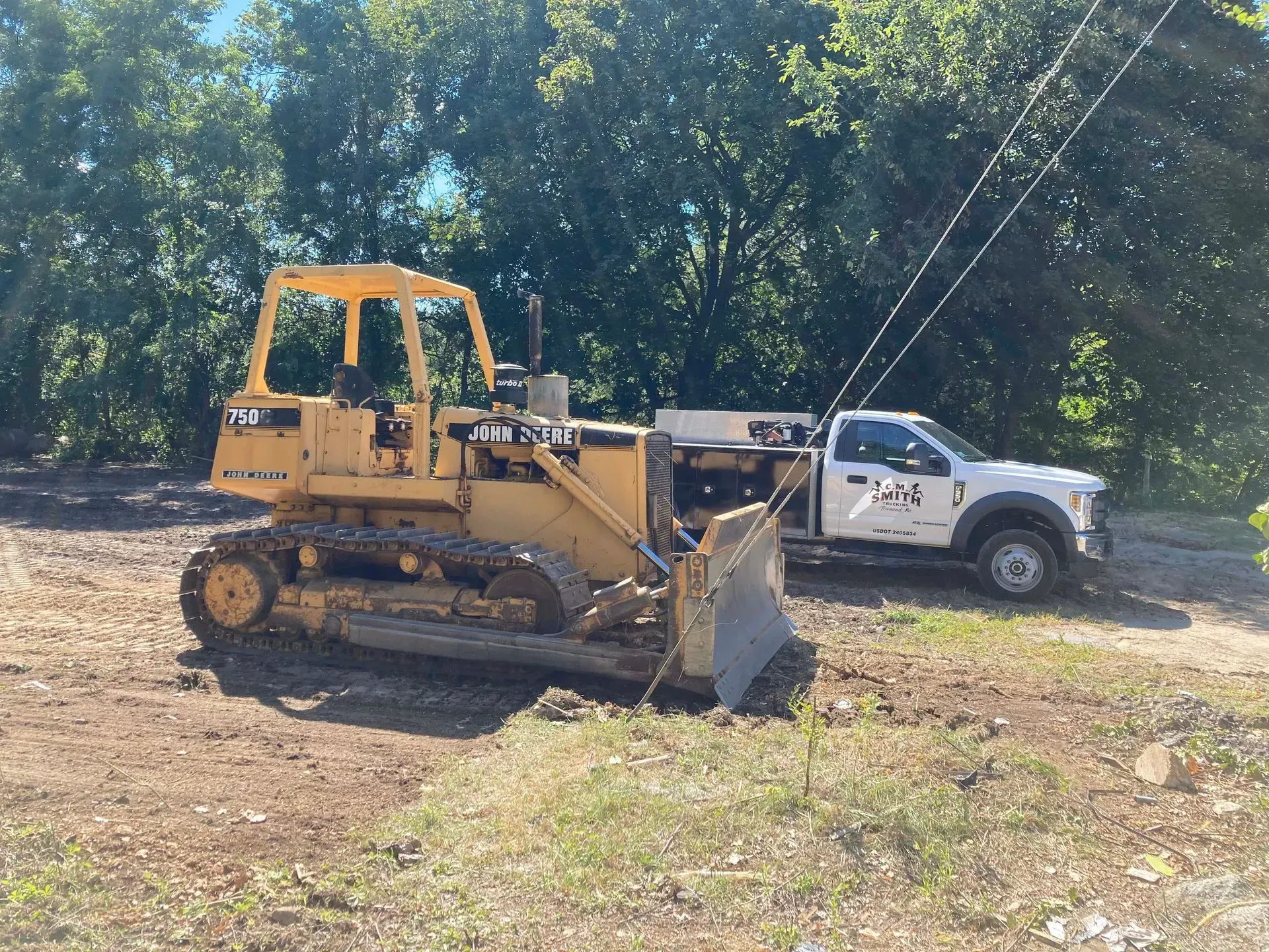 Yellow bulldozer beside a white pickup truck on a dirt clearing with trees in the background