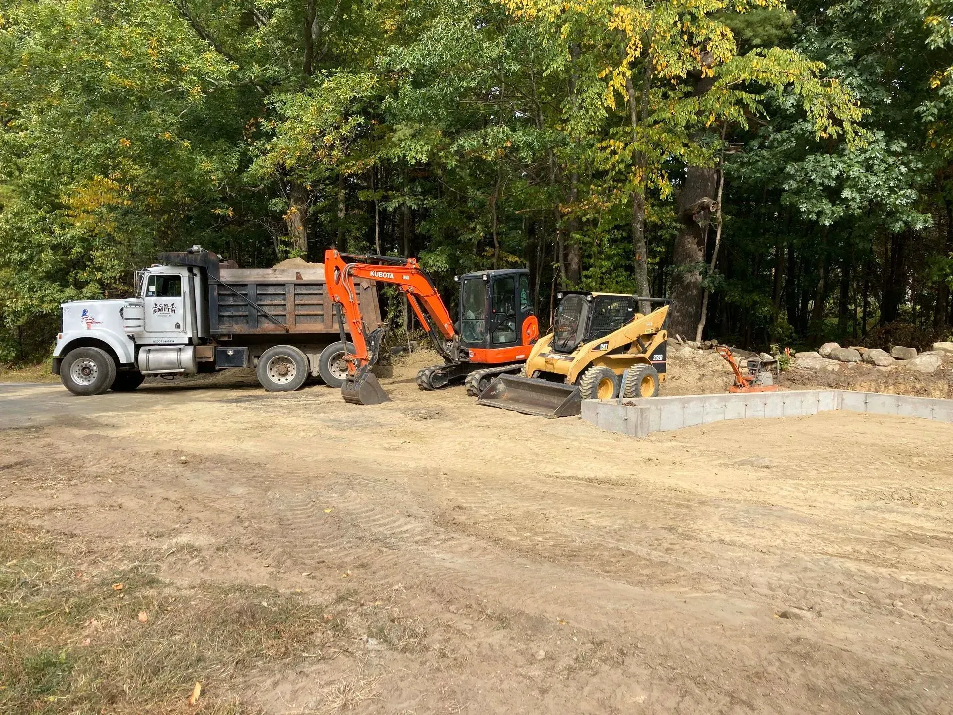 Dump truck, orange excavator, and yellow skid steer at a wooded construction site.