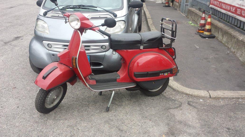 Uno scooter Vespa rosso parcheggiato su una superficie ghiaiosa, vicino a una piccola auto grigia e a un edificio.
