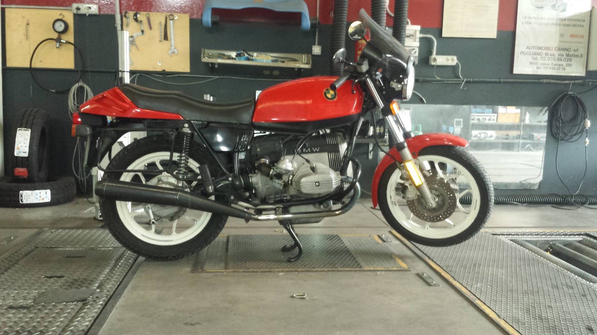 Moto rossa in un garage, su un ponte sollevatore. Cerchi bianchi e neri, motore argentato.