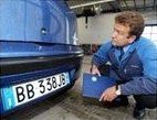 Un uomo esamina la targa di un'auto in un garage. Indossa un'uniforme blu e tiene in mano un blocco per appunti blu.