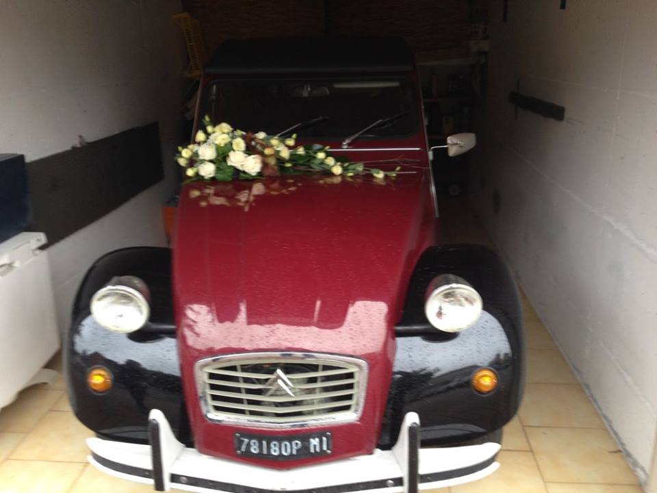 Una Citroën 2CV d'epoca, rossa scura e nera, decorata con fiori bianchi, parcheggiata in uno spazio stretto.