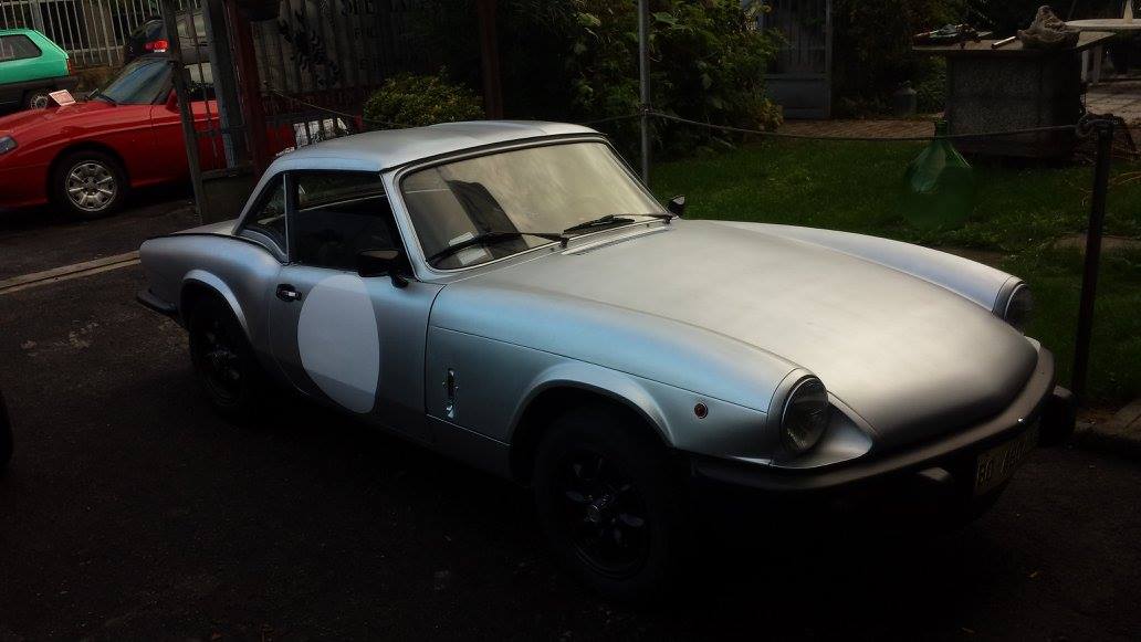 Auto sportiva Triumph Spitfire argentata con stemmi da corsa, parcheggiata all'aperto.